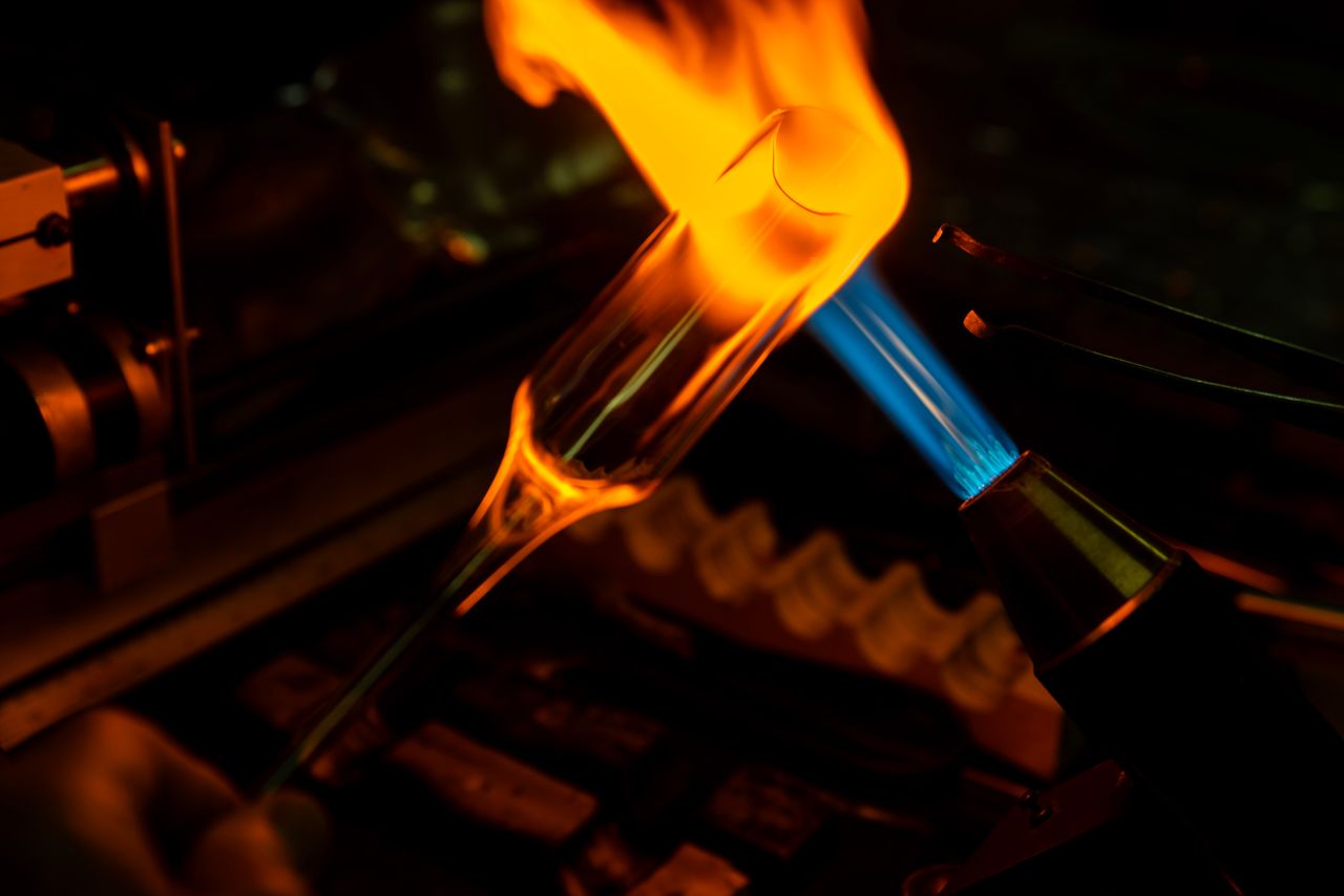 A close-up photograph of the glassblowing process with intense flames engulfing the glass.